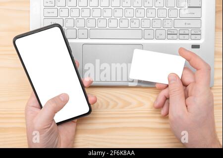 Achat en ligne. Homme mains tenir téléphone cellulaire avec écran blanc vierge et maquette de carte de crédit sur le fond de l'ordinateur portable. Homme tenant carte de crédit avec téléphone an Banque D'Images