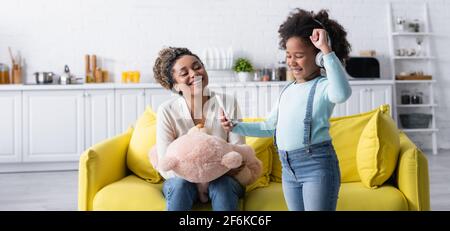 enfant afro-américain excité dans un casque montrant le geste de victoire près de la mère avec l'ours en peluche, bannière Banque D'Images