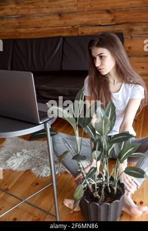 Pensive optimiste femme freelance élève dans une tenue décontractée assis sur le sol et regarder l'écran de l'ordinateur regarder le webinaire ou discussion vidéo Banque D'Images