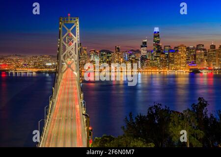 Bay Bridge avec des sentiers de lumière et le centre-ville de San Francisco vu de Treasure Island au coucher du soleil et des réflexions sur l'eau dans la baie, longue exposition Banque D'Images