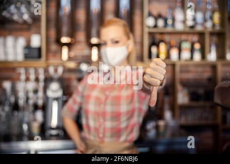 Travaillez dans un restaurant pendant le corona. Un portrait d'une serveuse féminine portant un masque avec une main sur la hanche tandis que l'autre main montre son pouce d Banque D'Images