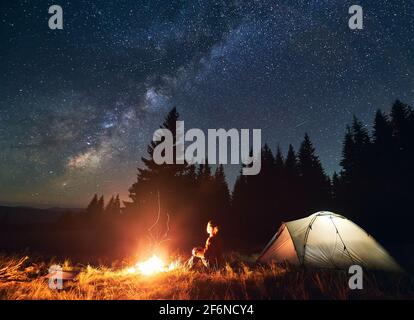 Camping de nuit dans les montagnes. Jeune randonneur assis près d'un feu de camp et d'une tente touristique illuminée. En arrière-plan forêt d'épinette sous beau ciel étoilé de nuit avec la voie lactée. Concept de tourisme Banque D'Images