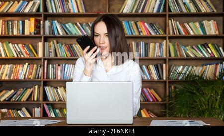 Bonne femme brune dans un élégant chemisier blanc parle sur mobile téléphone près d'un ordinateur portable contemporain à la table, contre de grandes étagères chambre Banque D'Images