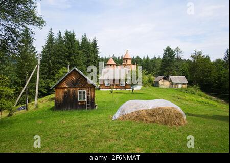 Maisons sur prairie verte dans un petit village et ancienne église en bois derrière. Tas de paille sèche. Paysage rural traditionnel dans les Carpathian Mountains. Ukraine. Banque D'Images