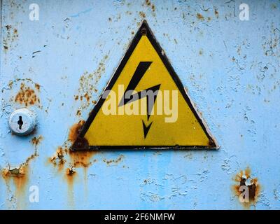 Ancien panneau haute tension. Triangle jaune sur une porte rouillée bleue. Symbole de danger. Banque D'Images