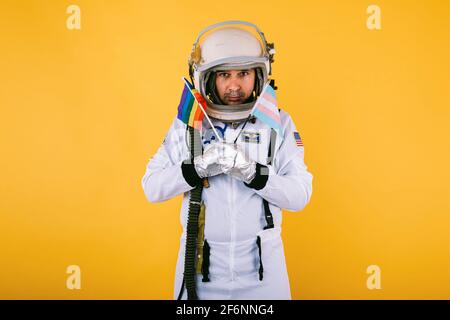 Cocosmonaute mâle transsexuel gay avec un geste sérieux dans la combinaison d'espace et le casque, tenant le drapeau arc-en-ciel lgtbi et le drapeau transgenre, sur fond jaune. Banque D'Images