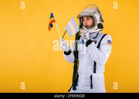 Cocosmonaute mâle transsexuel gay avec un geste sérieux dans la combinaison d'espace et le casque, tenant le drapeau arc-en-ciel lgtbi et le drapeau transgenre, sur fond jaune. Banque D'Images