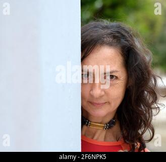 Sérieuse mature jolie femme caucasienne avec de longs poils peeks autour d'un mur gris et fait une expression faciale inquiète devant un fond de parc. Banque D'Images