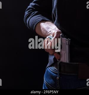 Main mâle prenant un pistolet de sa ceinture derrière le dos, sur fond noir. Banque D'Images