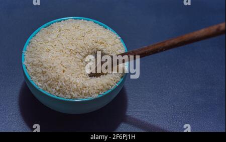 Riz cru servi dans une poêle à frire en porcelaine et une cuillère en bois sur fond sombre. Produit alimentaire humain. Nourriture naturelle. Grains de céréales éparpillés. Banque D'Images