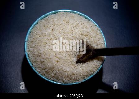 Riz cru servi dans une poêle à frire en porcelaine et une cuillère en bois sur fond sombre. Produit alimentaire humain. Nourriture naturelle. Grains de céréales éparpillés. Banque D'Images