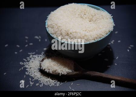 Riz cru servi dans une poêle à frire en porcelaine et une cuillère en bois sur fond sombre. Produit alimentaire humain. Nourriture naturelle. Grains de céréales éparpillés. Banque D'Images