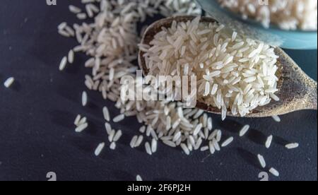 Riz cru servi dans une poêle à frire en porcelaine et une cuillère en bois sur fond sombre. Produit alimentaire humain. Nourriture naturelle. Grains de céréales éparpillés. Banque D'Images