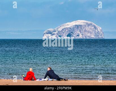 Un couple s'assoit sur une couverture de pique-nique sur la plage de Belhaven Bay avec vue sur la colonie de fous de Basso Rock, jour ensoleillé de la brise, East Lothian, Écosse, Royaume-Uni Banque D'Images