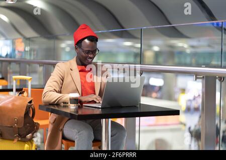 Homme de voyage africain assis à la table de café de l'aéroport, travaille à distance sur un ordinateur portable attendant un vol Banque D'Images