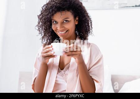 Femme afro-américaine en robe de soie tenant une tasse et souriant à l'appareil photo Banque D'Images