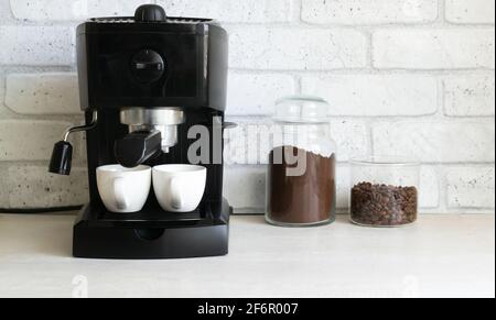 Une machine à café noire se trouve sur la table de cuisine, à côté de deux bocaux en verre avec du café moulu et des haricots. Le concept de faire du café à la maison. Hori Banque D'Images