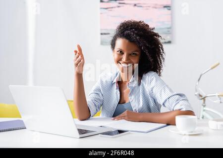 Femme afro-américaine pointant vers un appareil photo près de gadgets et d'une tasse de café sur un premier plan flou à la maison Banque D'Images