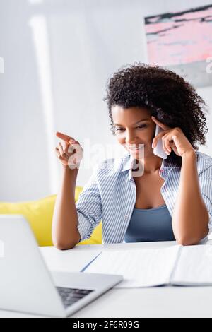 Femme afro-américaine pointant vers un ordinateur portable tout en parlant sur un téléphone portable près de l'ordinateur portable sur un premier plan flou Banque D'Images