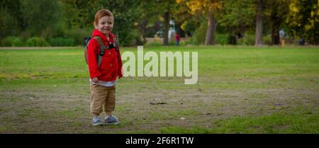 Un adorable petit garçon à tête rouge portant une veste rouge debout sur une pelouse dans un parc une soirée ensoleillée Banque D'Images