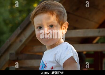 Portrait d'un adorable garçon à tête rouge et yeux bleus, portant un t-shirt blanc dans une aire de jeux par temps ensoleillé Banque D'Images