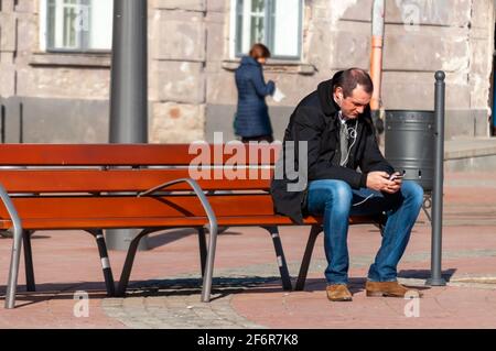 Timisoara, Roumanie - 14 mars 2016 : homme assis sur un banc dans la rue et écoutant de la musique depuis son téléphone. De vraies personnes. Banque D'Images