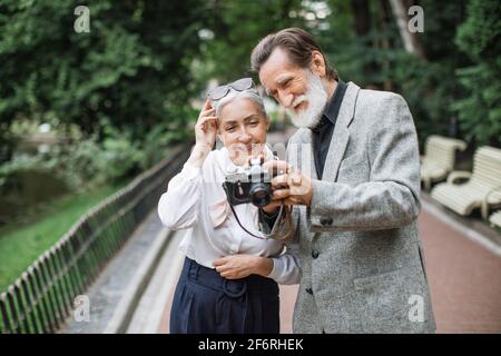 Homme et femme d'âge souriant debout dans un parc verdoyant et regardant des photos sur un appareil photo rétro. Couple d'âge mûr qui marche à l'extérieur et qui prend des photos. Banque D'Images