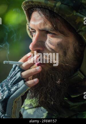 Homme de soldat avec sale visage fatigué dans le camouflage militaire et casque tenant une cigarette et fumant en plein air Banque D'Images