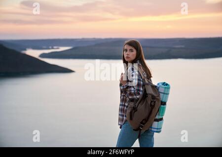 Charmante femme aux cheveux bruns regardant la caméra tout en se tenant debout au parc national Podillya Tovtry. Randonnée femelle avec sac à dos sur la nature pendant le coucher du soleil. Banque D'Images