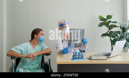 Une vieille femme médecin avec un écran facial et un masque de protection montre le film radiographique thoracique au patient assis dans un fauteuil roulant. Décrivez les symptômes o Banque D'Images