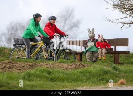 03 avril 2021, Bade-Wurtemberg, Dürmentingen : deux cyclistes féminins longent une ruelle de campagne, devant un banc sur lequel deux personnages de lapin de Pâques farcis de paille sont assis. Photo: Thomas Warnack/dpa Banque D'Images