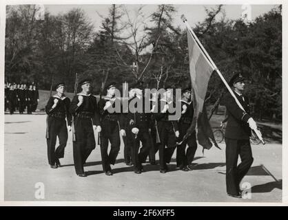 La photographie montre un groupe de jeunes recrues qui vont en parade et portent des fusils sur l'axe droit. Juste en relation avec le groupe il y a aussi une commande. Le groupe est situé au centre des images de l'image et devant elles, au premier plan à droite, se trouve également un support de ventilateur. Il tient la barre du ventilateur avec la main droite. La barre de ventilateur est couronnée de Gustav V Monogram et la bannière est un drapeau suédois à trois réglages. Tous ont leurs uniformes respectifs, complétés par des casquettes et des gants. En arrière-plan, on voit un groupe de recrues, de la végétation et de l'environnement du parc. La photographie Banque D'Images