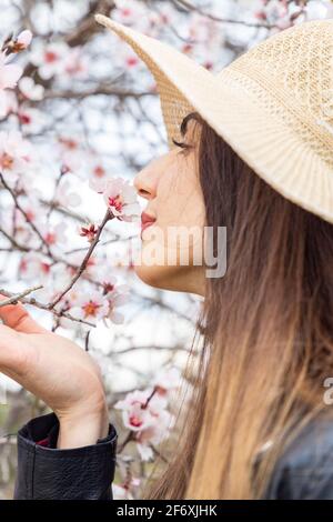 Belle femme brune heureuse avec chapeau, appréciant le parfum des fleurs sous un amandier fraîchement fleuri, le jour ensoleillé du printemps. Banque D'Images