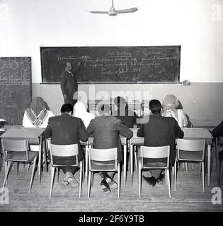 Années 1960, étudiants arabes historiques, certains portant des foulards de tête, assis dans une salle de classe avec un enseignant de sexe masculin au tableau noir, Djeddah, Arabie Saoudite. Il est intéressant de noter que la leçon semble être en anglais, car l'écriture sur le tableau noir sur les 'antibiotiques' est en anglais. Banque D'Images