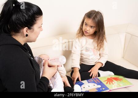 Fille de quatre ans regardant le livre d'images et parlant avec la mère, qui nourrit bébé soeur de 2 mois Banque D'Images