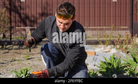 Recentrer les jeunes travailleurs de la construction dans des lunettes jaunes en éliminant les irrégularités sur la table de plancher à l'aide d'un marteau et d'un burin. Mains mâles en gants de travail rouges smas Banque D'Images