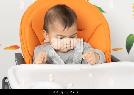 Petit garçon de six mois assis dans une chaise haute utiliser la pince pour prendre le snack de la chaise haute plateau Banque D'Images