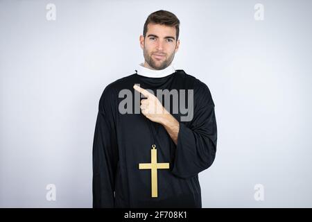 Jeune homme hispanique portant l'uniforme d'un prêtre debout sur fond blanc confus et pointant avec la main et le doigt sur le côté Banque D'Images