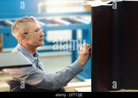 Un travailleur âgé restaure soigneusement les meubles dans un atelier de menuisier pendant la journée. Un charpentier moustachié avec des lunettes peint sur les rayures sur un meuble en bois. Banque D'Images