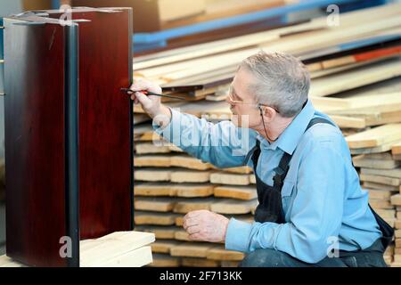 Un homme âgé avec des lunettes et des cheveux gris travaille avec du bois dans une boutique de menuiserie. Un travailleur d'apparence caucasienne est engagé dans la restauration de meubles. Emploi à temps partiel pour un pensionné Banque D'Images