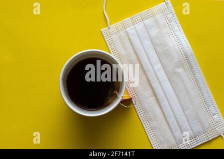 Une tasse blanche jetable remplie de thé, à côté d'un masque chirurgical jetable, fond jaune Banque D'Images