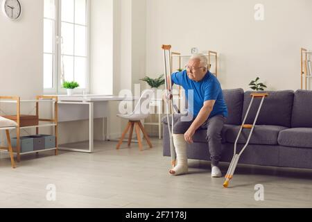 Homme mûr avec une jambe cassée essayant de se lever de canapé et marche avec béquilles à la maison Banque D'Images