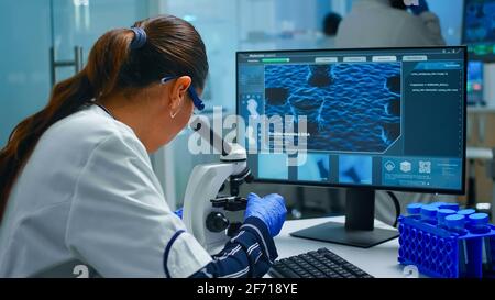 Biochimiste utilisant le microscope et l'écriture à l'ordinateur assis dans le laboratoire équipé. Médecin scientifique travaillant avec diverses bactéries, tissus, échantillons de sang, recherche pharmaceutique pour les antibiotiques Banque D'Images