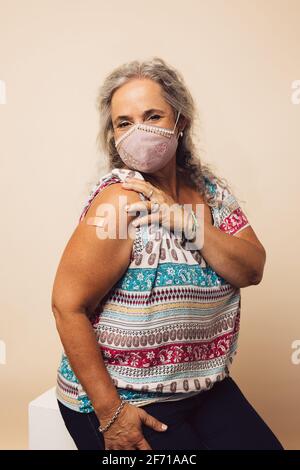 Femme âgée portant un masque facial montrant son épaule avec bandage après avoir reçu le vaccin. Femme mature assise sur fond marron après réception Banque D'Images