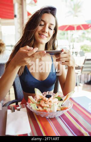 Jeune femme photographiant sa salade avec un smartphone tout en étant assise dans un restaurant Banque D'Images