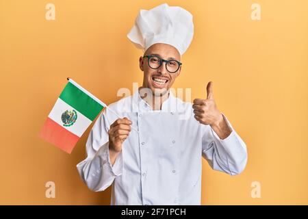 Homme chauve avec barbe portant un tablier de cuisinier professionnel tenant le drapeau du mexique souriant heureux et positif, pouce vers le haut faisant excellent et signe d'approbation Banque D'Images