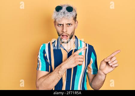 Jeune homme hispanique avec des cheveux teints modernes pointant avec les doigts sur le côté dans le visage de choc, regardant sceptique et sarcastique, surpris par la bouche ouverte Banque D'Images