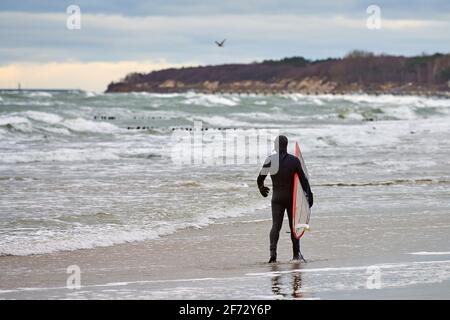 Surfeur masculin en maillot de bain noir marchant le long de la mer et tenant le surf blanc dans sa main. Journée chaude, beau ciel nuageux, scène de la nature. Sport et outdo Banque D'Images