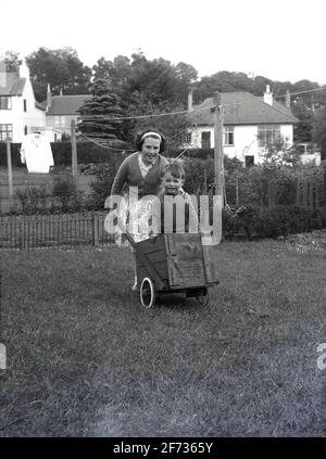 1956, historique, à l'extérieur dans un jardin à l'arrière, un petit garçon debout dans une charrette à deux roues en bois faite à la main étant poussé sur l'herbe par sa sœur, Dumfries, Soctland. Banque D'Images