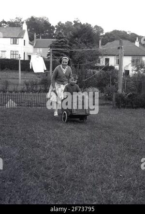 1956, historique, à l'extérieur dans un jardin à l'arrière, un petit garçon debout dans une charrette en bois faite à la main étant poussé sur l'herbe par sa sœur aînée, Dumfries, Soctland. Banque D'Images